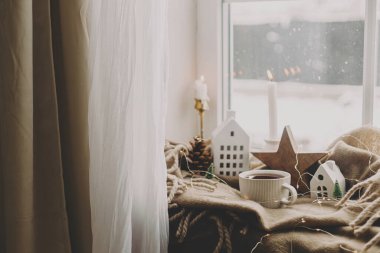 Sıcak bir fincan çay, mumlar, ışıklar, küçük bir noel evi ve ağaç pencere kenarındaki battaniyedeki ahşap yıldız. Kış kızarıklığı. Atmosferik Noel arkaplanı. Karlı bir günde sıcak bir ev.