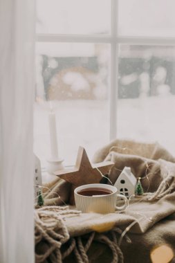 Kış kızarıklığı. Sıcak bir fincan çay, mumlar, ışıklar, küçük noel evleri ve ağaçlar, pencere kenarındaki battaniyenin üzerindeki ahşap yıldız. Rahat bir ev. Atmosferik karlı bir gün. Noel arkaplanı