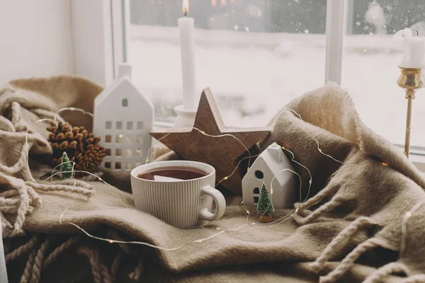 冬の寒さ キャンドル ライト 小さなクリスマスの家や木の暖かいカップ 窓辺の居心地の良い毛布の上に木製の星 居心地の良い家 雪の日だ クリスマスの背景 — ストック写真
