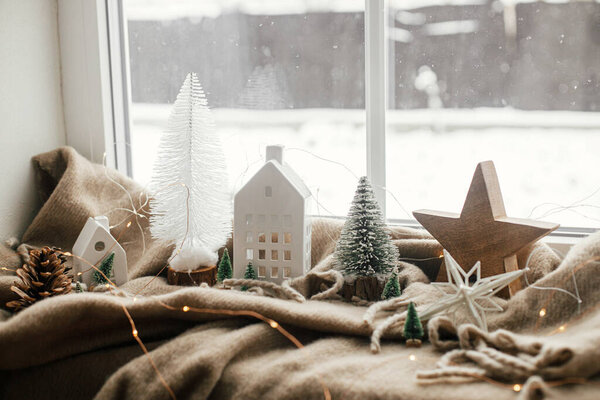 Christmas still life, winter hygge home. Stylish christmas decorations, tree, lights, little house and wooden star on cozy blanket on windowsill. Merry Christmas! Atmospheric scandinavian mood