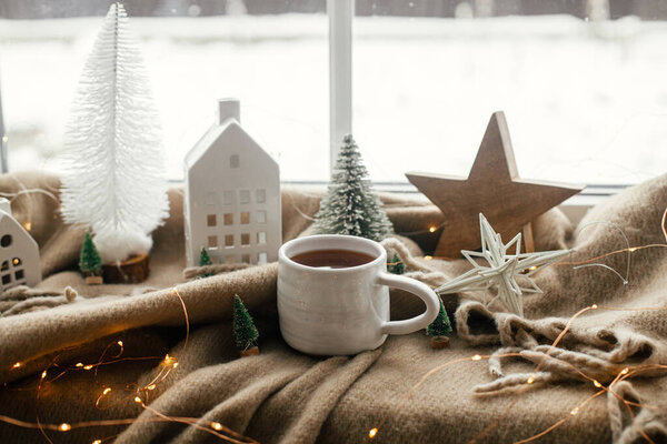 Christmas still life, winter hygge home. Warm cup of tea, christmas decorations, lights, little house, star on cozy blanket on windowsill. Cozy home. Atmospheric scandinavian mood