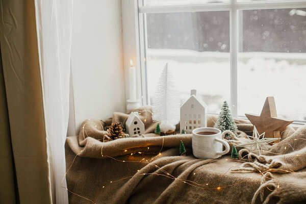 Winter hygge, Christmas still life. Warm cup of tea, christmas decorations, lights, little house, star on cozy blanket on windowsill. Cozy home on snowy day. Atmospheric scandinavian mood