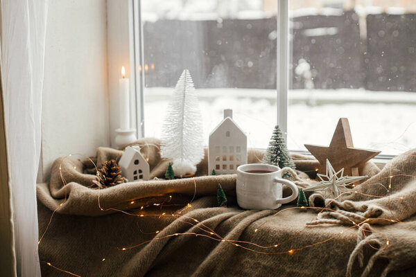 Warm cup of tea, christmas decorations, lights, little house, star on cozy blanket on windowsill. Winter hygge, Christmas still life. Cozy home on snowy day. Atmospheric scandinavian mood