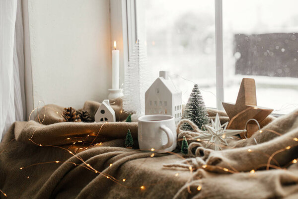 Winter hygge, Christmas still life. Warm cup of tea, christmas decorations, lights, little house, star on cozy blanket on windowsill. Cozy home on snowy day. Atmospheric scandinavian mood