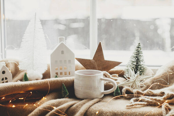 Christmas still life, winter hygge home. Warm cup of tea, christmas decorations, lights, little house, star on cozy blanket on windowsill. Cozy home. Atmospheric scandinavian mood