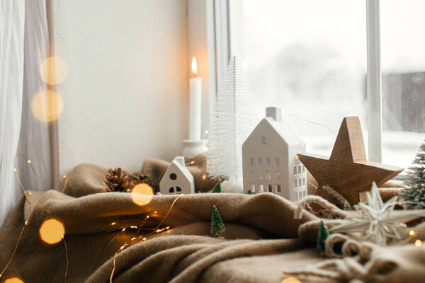 Winter hygge. Stylish christmas tree, lights, little house and wooden star on cozy blanket on windowsill. Merry Christmas! Atmospheric scandinavian mood, cozy home