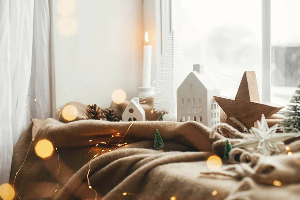 Christmas still life, cozy winter home. Stylish christmas decorations, tree, lights, little house and wooden star on cozy blanket on windowsill. Merry Christmas! Atmospheric scandinavian mood