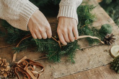 Kadife kurdele tutan ve kırsal ahşap masada Noel çelengi süsleyen kadın eller. Noel kırsal çelengi yapıyorum. Huysuz bir görüntü. Kış tatili atölyesi, Noel hazırlıkları