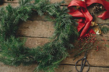 Noel çelengi yapıyorum. Firavun dalları yuvarlak Noel çelengi, kırmızı kurdele, böğürtlen, kırsal ahşap masada makas. Huysuz tatil imajı. Tatil atölyesi, kış dekorasyonu.