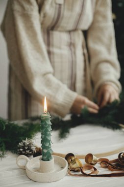 Modern Noel çelengi yapıyorum. Süslü mum köknar ağacı gibi, altın çanlar ve kurdele arka planda beyaz ahşap masada boho çelengi yapan kadın elleri. Kış dekoru, karamsar görüntü
