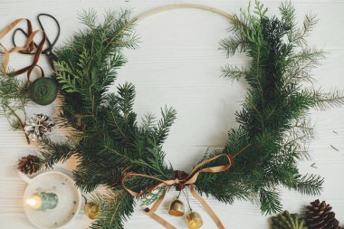 Modern Noel çelengi düzdü. Kadife kurdeleli ve altın zilli şık Noel çelengi, mum, çam kozalağı, beyaz ahşap masada makas. Tatil dekoru, karamsar bir görüntü. Mutlu Noeller.!