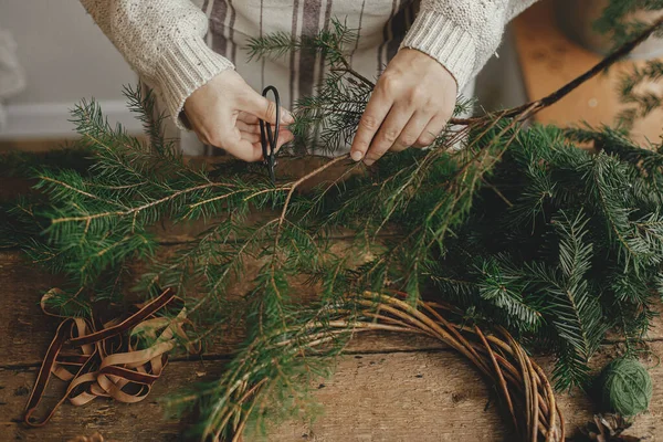 Making rustic Christmas wreath. Female hands cutting fir branches with scissors on background of rustic wooden table with ribbon. Moody Christmas image, holiday preparations