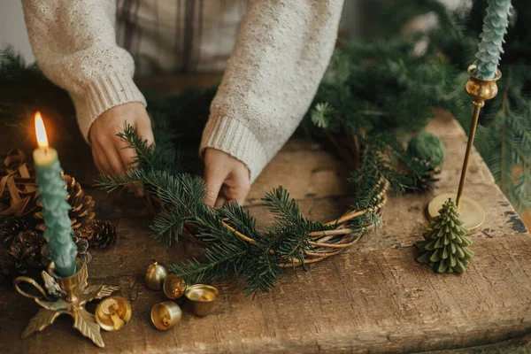 クリスマス素朴な花輪 モミの枝を保持し リボン 黄金の鐘 ろうそくと素朴な木製のテーブルの上に花輪を作る女性の手 ムーディークリスマスのイメージ休日の準備 — ストック写真