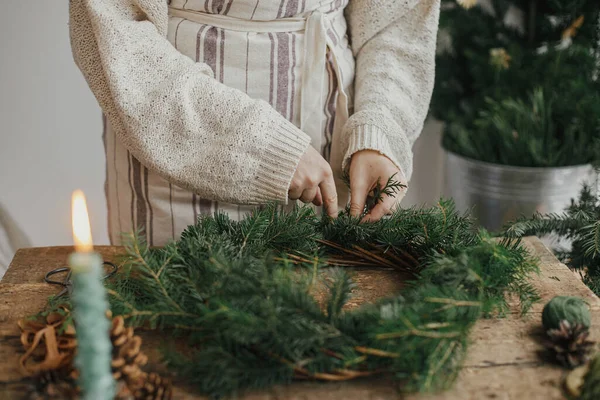 让圣诞乡村花环 女人的手拿着香柏木树枝 金色铃铛 蜡烛在乡村木桌上做花圈 喜怒无常的假日形象 寒假工作坊 — 图库照片