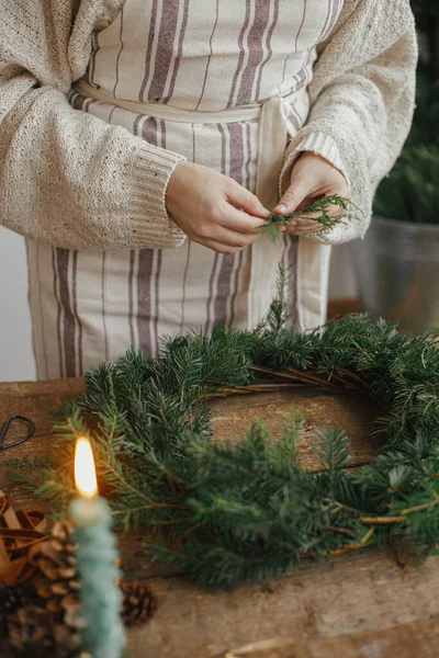 クリスマスの素朴な花輪を作る 女性の手は杉の枝を保持し リボン 黄金の鐘 ろうそくと素朴な木製のテーブルの上に花輪を作る ムーディーの休日のイメージ 冬休みワークショップ — ストック写真