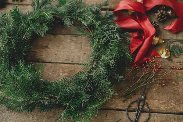クリスマスの花輪を作る ラウンドクリスマスリース 赤いリボン ベリー 素朴な木製のテーブルのはさみで公正な枝 ムーディーの休日のイメージ 休日のワークショップ 冬の装飾 — ストック写真