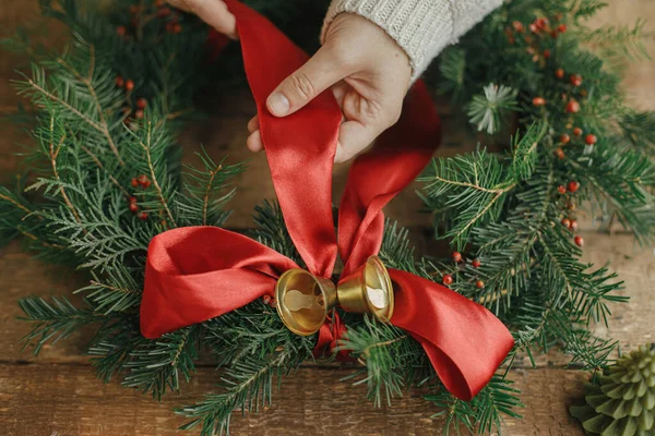 松のコーンとろうそくと素朴な木製のテーブルの上に赤いリボンと黄金の鐘でクリスマスリースを飾る女性の手 クリスマスの花輪 気分の良い休日のイメージを作る 休日ワークショップ — ストック写真