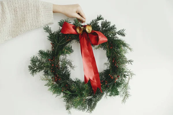 Merry Christmas and Happy Holidays! Hand holding stylish christmas wreath with red ribbon and golden bells on white wall background, moody image. Winter holiday decor
