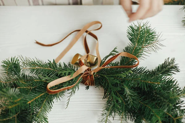 クリスマスの現代的な花輪を作る 白い木製のテーブルの上に黄金の鐘やリボンで花輪を飾る女性の手 休日のワークショップ 冬の装飾 ムーディなイメージ — ストック写真
