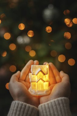 Merry Christmas and Happy Holidays! Little glowing house in hands on background of illuminated christmas tree lights bokeh. Magical time. Cozy home, atmospheric image. Family and safety concept clipart