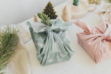 Sıfır Noel konsepti. Beyaz köy masasında keten kumaşlara sarılmış şık hediyeler. Çevre dostu ahşap oyuncaklar, köknar dalları, mum. Furoshiki hediye paketi. İyi tatiller.!