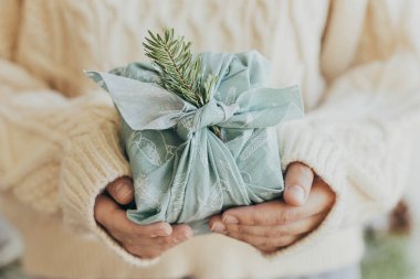 Mutlu Noeller ve mutlu tatiller! Yeşil kumaşa sarılı, köknar dallı bir hediye. Şık Furoshiki hediyesi. Örgü kazaklı bir kadın çevre dostu bir hediye tutuyor. Sıfır kayıp Noel