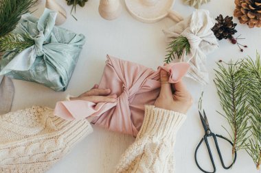 Beyaz ahşap masa üzerinde pembe keten kumaşla hediye paketi, köknar dalı, çevre dostu oyuncak, mum. Sıfır Noel konsepti. Furoshiki hediye paketi, üst görünüm