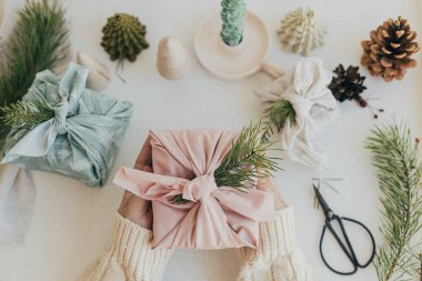 Sıfır atık Noel yatıyordu. Furoshiki hediye paketi. Elleri pembe kumaşa sarılı, köknar dallı hediye paketi beyaz ahşap masa üzerinde, çevre dostu dekor ve mum ile. Mutlu Noeller.!