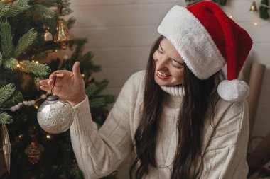 Mutlu Noeller! Rahat süveter ve Noel Baba şapkalı mutlu bir kadın. Atmosferik festival odasında şık takılarla Noel ağacı süslüyor. Kış tatili hazırlığı