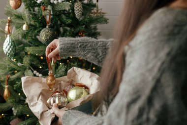 Rahat süveter giymiş şık bir kadın. Atmosferik şenlik odasında şık bir takı ile Noel ağacı süslüyor. Mutlu Noeller! Kış tatili hazırlığı. Xmas ağacını eski oyuncakla süslemek