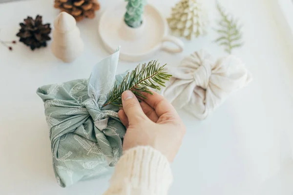 stock image Zero waste Christmas concept. Hands decorating gift box wrapped in fabric with fir branch on white wooden table with eco friendly decor and candle. Furoshiki gift wrapping. Eco winter holiday