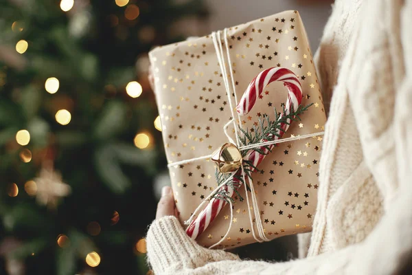 ライト付きクリスマスツリーに手をつないでスタイリッシュなクリスマスプレゼント メリークリスマスとハッピーホリデー 女性で居心地の良いセーター保持ラッププレゼントとともにキャンディー杖で大気中の部屋 — ストック写真