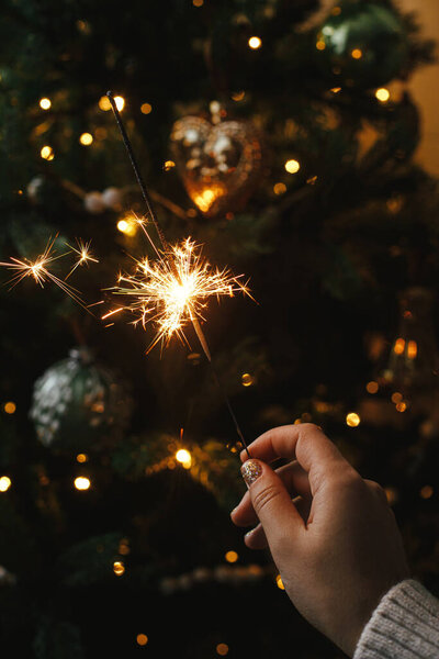 Горящий блеск в женской руке на фоне рождественских ёлочных огней в темной комнате. С Новым Годом! Рука держит фейерверк против стильного украшенного дерева с подсветкой. Атмосферное время
