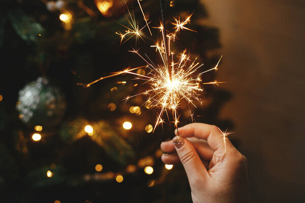 Happy New Year! Burning sparkler in female hand on background of christmas tree lights in dark room. Atmospheric celebration. Hand holding firework against stylish decorated tree with illumination
