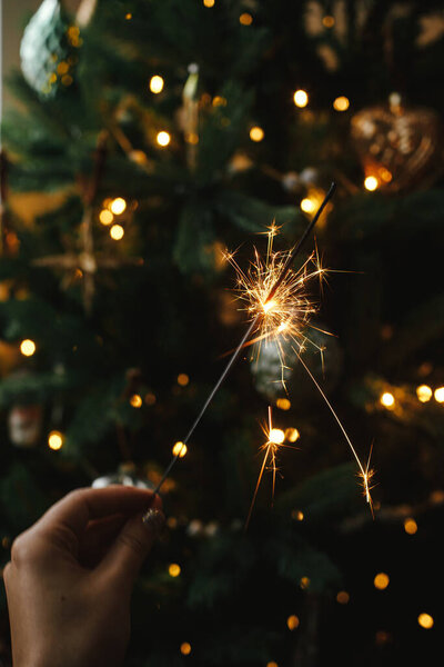 Hand holding firework against christmas tree lights in dark room. Happy New Year! Merry Christmas! Burning sparkler in female hand  on background of golden illumination bokeh. Atmospheric time