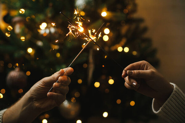 Hands holding firework against christmas tree lights in dark room. Happy New Year! Couple celebrating with burning sparklers in hands on background of stylish decorated tree with illumination.
