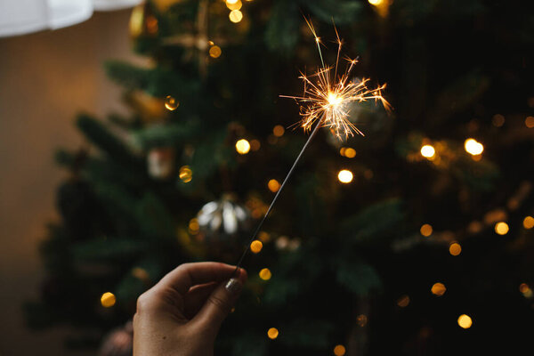 Рука держит фейерверк против рождественских огней в темной комнате. С Новым Годом! Счастливого Рождества! Горящий бенгальский огонь в женской руке на фоне золотого освещения боке. Атмосферное время