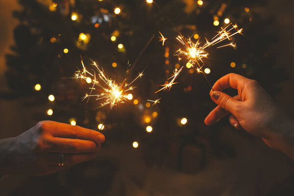 С Новым Годом! Пара празднует с горящими искрами в руках на фоне стильного декорированного дерева с подсветкой. Руки держат фейерверк на рождественских ёлочках в темной комнате