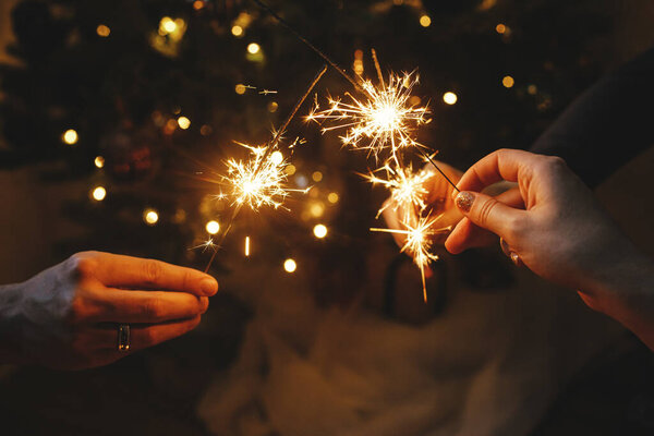 Руки держат фейерверки против рождественских огней в темной комнате. С Новым Годом! Атмосферный праздник. Друзья, празднующие с горящими бенгальскими огнями в руках на фоне стильного освещенного дерева