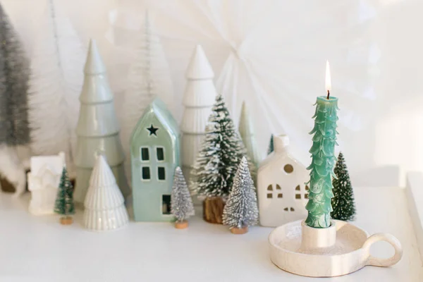 Stylish christmas tree candle on background of winter miniature snowy village on white table. Modern little Christmas trees and houses. Holiday advent. Merry Christmas and Happy Holidays!
