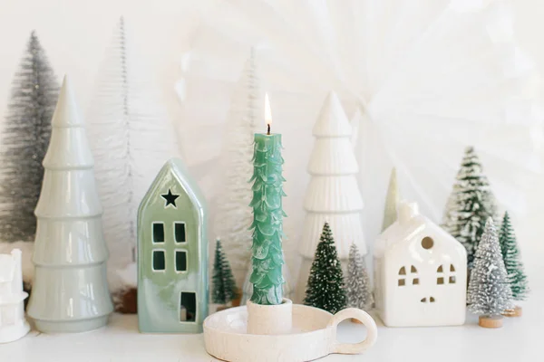 Merry Christmas and Happy Holidays! Stylish christmas tree candle on background of winter miniature snowy village on white table. Modern little Christmas trees and houses. Holiday advent
