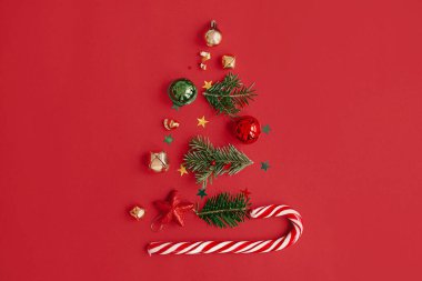 Noel ağacı, kış tatili konsepti. Kırmızı arka planda köknar dalları, mücevherler, altın konfeti ve şeker kamışından yapılmış şık bir Noel ağacı, düz bir zemin. Yaratıcı bir fikir. Modern bayrak