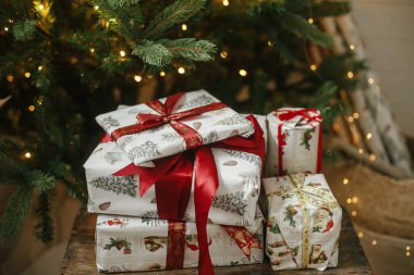 Işıklı Noel ağacında şık bir Noel hediyesi. Mutlu Noeller ve mutlu tatiller! Karamsar odada süslenmiş ağacın altında kırmızı kurdeleyle paketlenmiş Noel hediyeleri. Atmosferik pankart