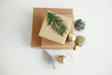 Beyaz masa üzerinde şık eko-noel hediye kutuları vardı. Basit el sanatları hediyeleri altın takılar, kırsal ahşap masa üzerinde çan ve köknar dalı. Mesaj için yer var. Mutlu Noeller.