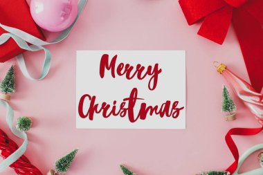 Üzerinde kırmızı fiyonk, küçük ağaçlar, mücevherler, pembe arka planda düz kurdeleler olan mutlu noel mesajları. Sezonun tebrik kartı. El yazısı işareti