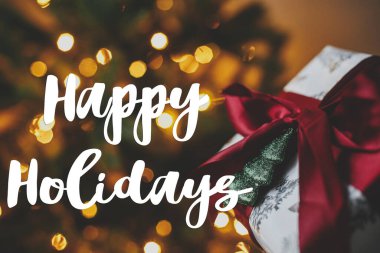 Noel ağacının arka planında, akşam odasında altın ışıklarla süslenmiş şık bir Noel hediyesi üzerine Mutlu Tatiller tabelası. Sezonun tebrik kartı. Atmosferik istekler