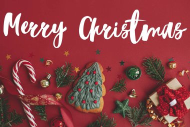 Noel ağacı kurabiyesi, hediye, takılar, köknar dalları, kırmızı arka plandaki konfeti üzerine Mutlu Noeller mesajı. Sezonun tebrik kartı. El yazısı işareti