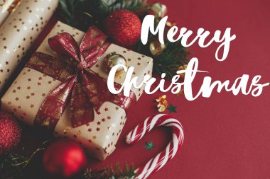 Noel hediyesi, ambalaj kağıdı, köknar dalları, şeker kamışı ve kırmızı arka planda takılar üzerine Mutlu Noeller metni. Sezonun tebrik kartı. El yazısı işareti