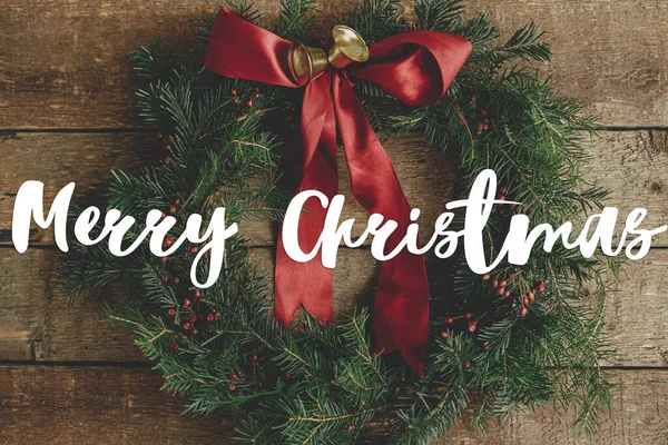 圣诞快乐的圣诞祝福 用红丝带 浆果和金色的铃铛装饰着时髦的圣诞花环 躺在质朴的木桌上 季节的贺卡 手写签名 — 图库照片