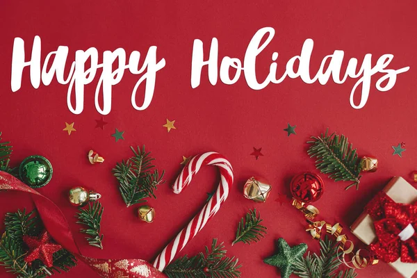 幸せな休日のテキストサイン上のクリスマスギフト モミの枝 コンフェッティとキャンディの杖赤の背景フラットレイアウト シーズンのグリーティングカード クリスマスポストカード — ストック写真